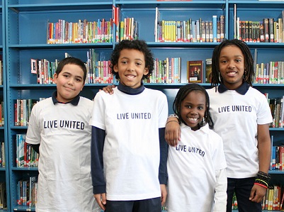 4 children with short brown hair, brown eyes wearing white t-shirts that have black lettering that says Live United on the front. Bookcase behind them with blue background.