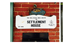 Meeting Ground House Logo with red colored brick background and white sign with black lettering that says Settlement House in uppercase letters. There is a blue welcome sign written in black lettering on top of the white sign.