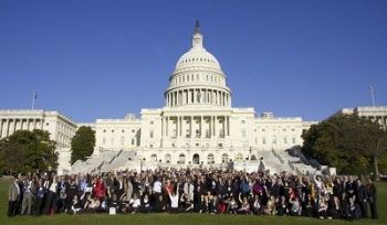 White House Building in white with blue background with many people posing for a photo. Green Trees in the background.